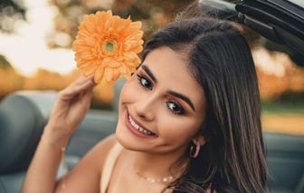 A beautiful Latina in a yellow dress sitting in a convertible car holding a flower by her right ear.
